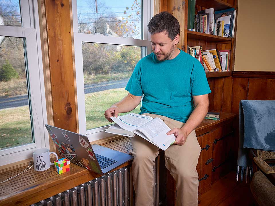male student reading textbook while sitting near window