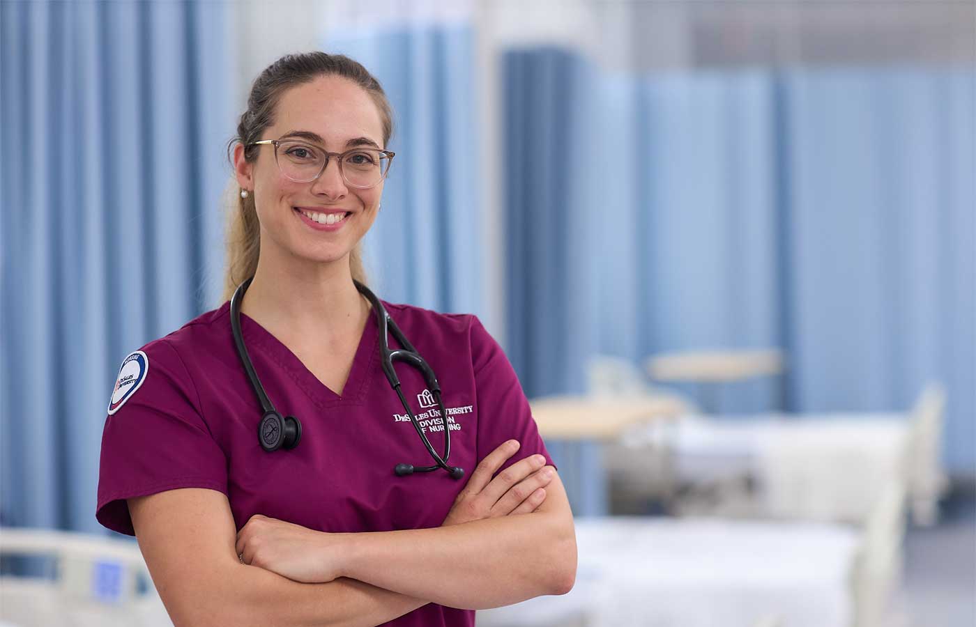 A nursing student at DeSales University smiles confidently in a clinical lab setting, wearing a maroon uniform and stethoscope